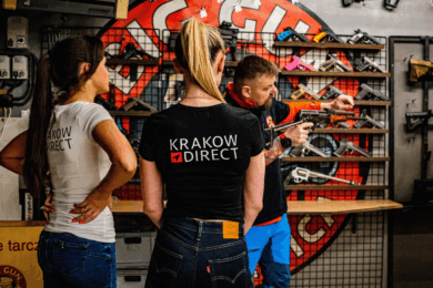 Instructor briefing two women at a Krakow shooting range, with various firearms displayed on the wall. Both women wear "Krakow Direct" branded shirts